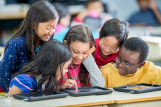 A multi-ethic group of elementary age children are sitting in the computer lab and are looking at a tablet together.